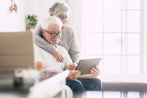An elderly man is sitting on a couch with a woman, and they are both smiling while looking at a tablet