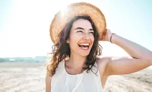 A woman smiling on a beach with a hat on and the sun shining brightly behind her