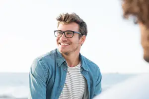 A man with glasses smiling at another person on the beach.