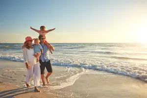 A family is walking on the beach during sunset. The man is carrying a child on his shoulders, and the woman is holding a child's hand.