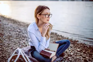 Woman with glasses reading a book by the lake at sunset