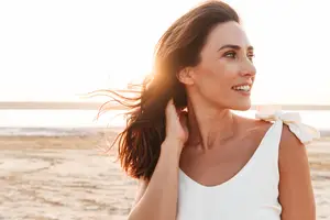 A woman with long hair blowing in the wind, smiling at the camera on a beach.