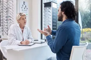 A man and woman are seated at a desk, possibly discussing eyeglasses, with the woman taking notes and the man gesturing. Behind them, there is a large window with a view of a garden and a vision chart on the wall.
