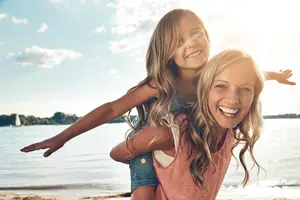 Mother carrying her daughter on her back at the beach on a sunny day.