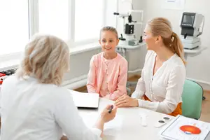 Two women are having a discussion with a girl in an office setting, possibly a doctor's office. The woman on the left is holding a pen and paper, and the woman on the right is smiling at the girl. The girl is wearing a pink shirt and is sitting in front of a desk with a monitor and other medical equipment. There is a window behind them with a view of the outside.