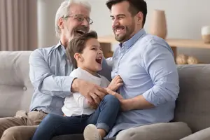 A man and his son are sitting on a couch laughing together, while the father holds the child's hand.