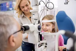 A young girl is having her eyes examined by an optometrist while an adult woman and a man look on in an optical clinic.