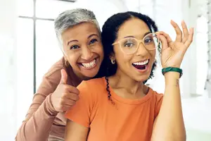 Two women are smiling and posing for a picture. One is wearing glasses, and the other has her hand raised, giving a thumbs-up sign.