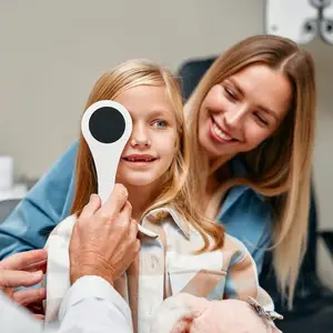 A young girl is having her eyes checked by a doctor while a woman holds her and smiles
