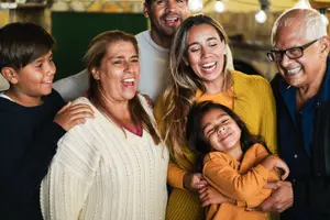 A family of five, including an elderly man, a woman, a girl, a boy, and a man, are smiling and posing for a photo in a room with a brick wall and a green door, along with some hanging lamps and shelves.