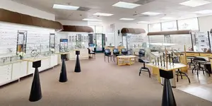 Interior of an optometrist office with chairs, tables, and display cases with eyewear. The room has a warm color scheme with beige walls and brown furniture. There are lights on the ceiling and large windows that let in natural light. The display cases are filled with various styles of glasses and accessories.