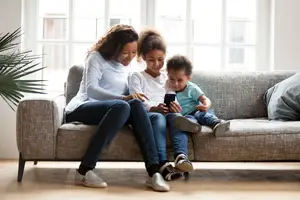 A family of three, two girls and a boy, sitting on a couch, looking at a cell phone