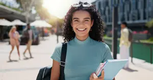 A woman in a blue turtleneck holding a folder and books, smiling while walking on a sidewalk with people in the background