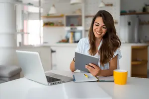 Woman sitting at a table in a kitchen with a laptop and tablet smiling at the camera