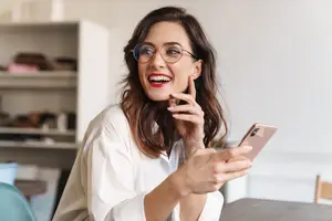 A woman with glasses is smiling while holding a cell phone in her right hand and touching her chin with her left hand.