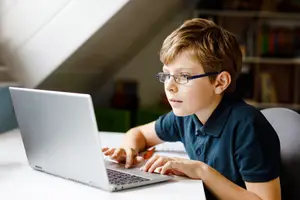 A young boy wearing glasses is using a laptop while sitting on a chair in a room with a shelf and a desk.