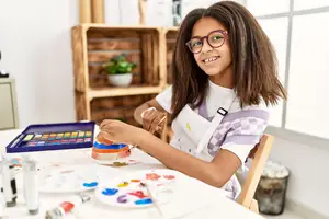 A girl wearing glasses and an apron sits at a table with a paint palette and brushes, smiling as she paints a cup.