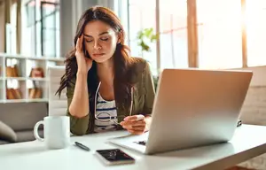 A woman sits at a desk with a laptop, cup, and book, appearing to be studying or working. The desk is cluttered, and she holds her head with her left hand.