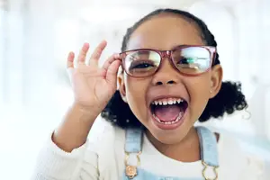 Close-up of a young girl with curly hair wearing glasses and smiling with her mouth open.
