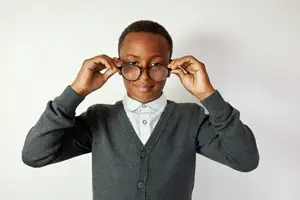 A young boy wearing glasses and a gray cardigan is smiling while holding the glasses up to his face.