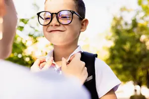 A boy wearing glasses and a backpack smiles while a person adjusts his collar in an outdoor setting.