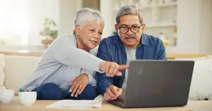 An elderly couple is sitting on a couch, pointing at a laptop on a table.