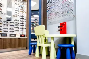 A kid's table and chairs are placed in a shop, with many glasses displayed on the wall and a sign that reads 'HUGO' on the wall. The shop has a blue door and a wooden counter. The shop has a wooden floor and a white wall. The shop has a bright light on the ceiling.