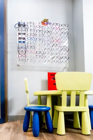 A room with a yellow table and chairs, a red book titled Hug, and a wall with a display of eyeglasses. The wall has a white background with a brown border, and the floor is wooden.