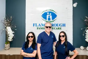 Three people in blue scrubs are standing in front of a white wall with a blue logo that says 'FLAM ROUGH FAMILY CARE'. The woman on the left has a ring on her finger and a bracelet on her wrist. The woman in the middle is wearing sunglasses and has a necklace. The man in the middle is also wearing sunglasses and a ring. There are two clocks on the right side of the wall, and two vases with white flowers on the left side.