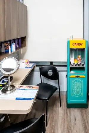 A room with a desk and chair, a candy machine, and a mirror. There are also cabinets with various items on them, and a window on the wall.