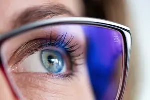 A close-up of a woman's eye with blue iris and eyelashes reflected in her glasses.