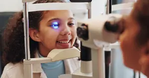 A young girl with curly hair is looking into an ophthalmoscope while a woman examines her eyes with a device, possibly in an eye clinic.