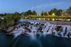 Water is flowing over rocks in a river with a city in the background.