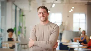 A smiling man wearing glasses and a beige long-sleeve shirt stands with his arms crossed in an office with other people sitting and working in the background.