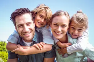 A family of four standing together smiling for a picture in a park with trees in the background