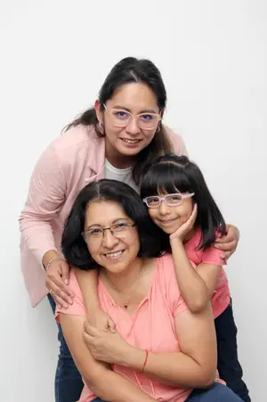 Three women are smiling for a photo, with the older woman wearing glasses and a necklace, the younger woman in a pink jacket, and the child wearing pink glasses.