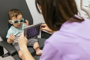 A child wearing sunglasses is sitting on a chair and holding a tablet in his hands while a woman is holding the tablet in front of him. The woman is probably a doctor, and she is checking the child's eyes with a tablet.
