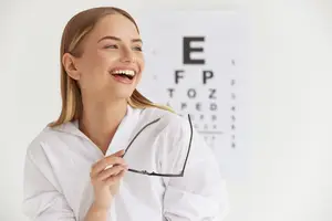 A smiling woman wearing glasses is holding a pair of glasses and seems to be posing for a photograph in a room with a white wall in the background.