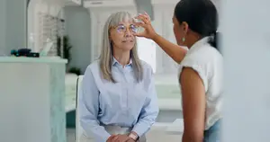 An older woman wearing glasses is having her vision tested by a woman in a white shirt in an eye clinic.