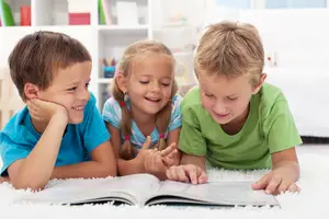 Three children are lying on a white carpet and reading a book together