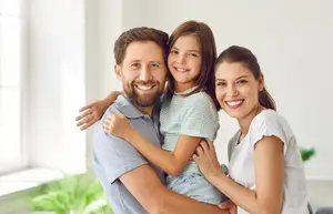 A family of three, consisting of a father, daughter, and mother, are posing for a photograph in a room with a white wall and a window with a white frame. The man is holding the girl, and the girl is holding the woman's hand, and they are all smiling for the camera.