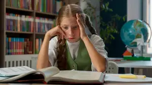A young girl is sitting at a desk in a classroom, looking stressed while holding her head with her hands. There is a globe and a house model on the desk in front of her. Behind her, there is a shelf with many books on it.