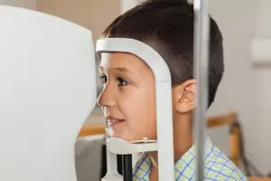 A young boy wearing a blue checkered shirt is getting an eye exam with a white machine in a room with white walls.