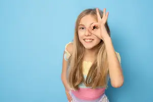 A young girl with long blonde hair is making a peace sign with her hand and smiling in front of a blue background
