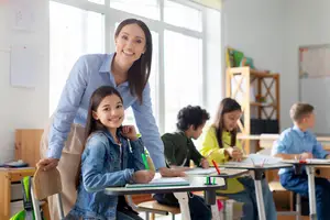 A woman teacher is smiling while teaching a student with a pencil in a classroom with other students sitting at their desks