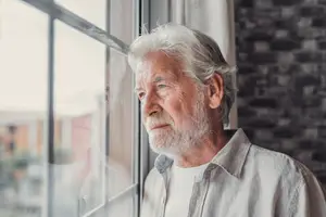 An elderly man with white hair and a beard is standing and looking out of a glass window with a white curtain on the side. He is wearing a white polo shirt and has a serious expression on his face. He is probably looking at something outside the window. Behind him is a blurry view of a brick wall and a building with a glass window.