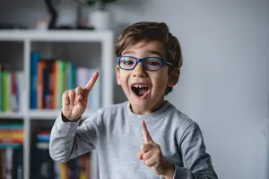 Happy boy with glasses pointing with both hands inside a room with shelves filled with books