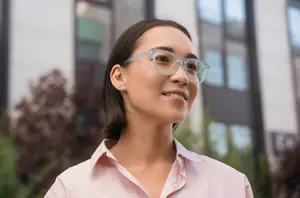 A woman wearing glasses is smiling in front of a building with glass windows and plants