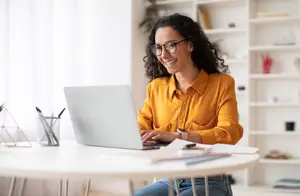 A smiling woman wearing glasses and a yellow shirt sitting at a desk with a laptop and other office supplies