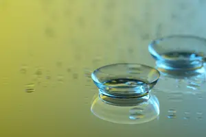 Two clear glass contact lenses are sitting on a yellow surface with small water droplets scattered around.